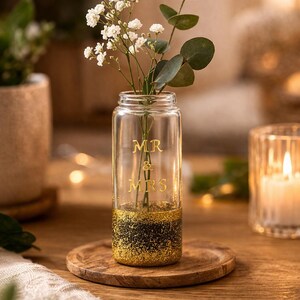 May include: A clear glass vase with gold glitter at the base and the words "MR & MRS" in gold lettering. The vase holds white flowers and greenery. It sits on a wooden coaster, with a lit candle in the background.