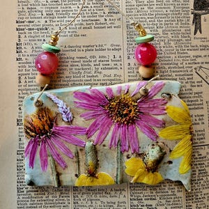 May include: A pair of earrings with pink, green, and white beads. The earrings are hanging from a ceramic tile with a floral design. The tile has a pink, yellow, and green flower design. The tile is attached to a page from a dictionary with the word "kit" highlighted.