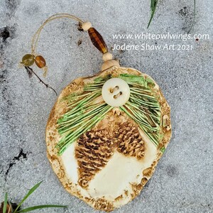 May include: A handmade ceramic ornament shaped like a pine cone with green and brown accents. The ornament has a white button and a loop for hanging. www.whiteowlwings.com Jodene Shaw Art 2021