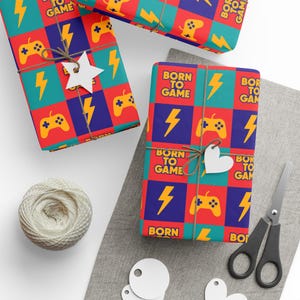 May include: Gift-wrapped presents with a colorful pattern featuring game controllers, lightning bolts, and the text "BORN TO GAME." The gifts are tied with twine and adorned with paper cutouts. A spool of twine and scissors are also present.
