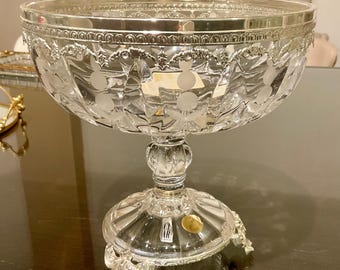 May include: A clear glass pedestal bowl featuring a silver-toned metal rim with an embossed pattern. The bowl has etched floral motifs along the sides, a thick glass stem, and a circular base supported by three ornate silver-toned metal feet.