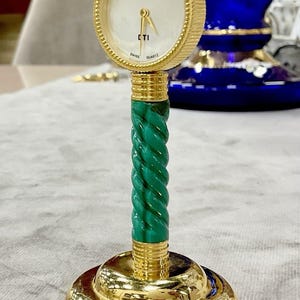 May include: A desk clock featuring a round white face with gold hands, a logo, and the text "DTI" and "SWISS QUARTZ". The clock is mounted on a green malachite-patterned twisted column with a polished gold-tone circular base and a ribbed gold bezel.