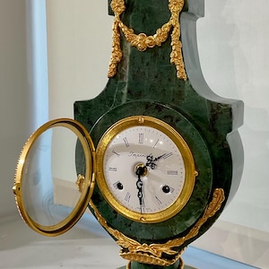 Op de afbeelding: Een donkergroene marmeren schoorsteenklok met een witte ronde wijzerplaat, zwarte Romeinse cijfers en de tekst Imperial. De klok is voorzien van goudkleurige metalen versieringen, waaronder een scharnierende glazen deur en decoratieve bloemenslingers.