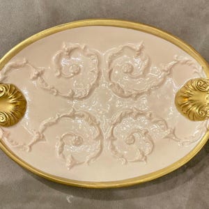 May include: An oval-shaped cream-colored ceramic tray featuring a raised relief scrollwork pattern across the surface. The tray is framed by a metallic gold rim and includes two decorative gold shell-shaped handles on the left and right sides.