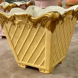 May include: A square ceramic planter with a pale yellow lattice pattern on the exterior and a white interior. The top rim features ornate, raised gold-colored scrollwork detailing. The base is supported by four small, square-shaped feet.