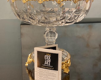 May include: A clear crystal bowl with a faceted pattern, featuring a gold-toned metal rim with ornate filigree detailing. The bowl rests on a clear crystal pedestal base with gold-toned metal feet. A white informational tag with the text CRE ART is attached.