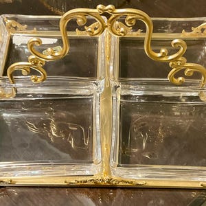 May include: A rectangular serving tray featuring four clear glass square compartments set within a gold-toned metal frame. The frame includes an ornate, curved gold handle centered between the glass sections, with decorative metal accents at the corners.