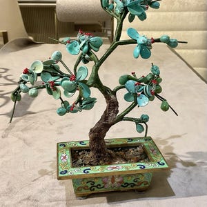 Puede incluir: Una escultura decorativa de árbol bonsái con un tronco marrón texturizado y ramas de alambre verde adornadas con pétalos de piedra azul claro y pequeños detalles de cuentas rojas. El árbol está en una maceta rectangular de metal.
