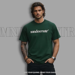 以下が含まれることがあります： 胸元に白い「mndovrmtr」の文字が入ったダークグリーンの半袖Tシャツ。クルーネックでゆったりとしたフィット感です。背景には大きな輪郭で「MND」と「TR」の文字があり、下部には「LIVE YOUR QUIRKS. WEAR YOUR CHAOS」と記載されています。