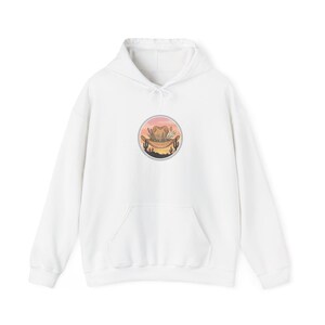 Könnte beinhalten: Ein weißer Kapuzenpullover mit einer kreisförmigen Grafik auf der Brust. Das Design zeigt einen braunen Cowboyhut, der mit Federn und Blättern verziert ist, vor einem Sonnenuntergangshimmel mit Silhouetten von Wüstenkakteen.