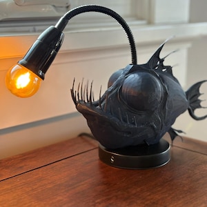 May include: A black anglerfish lamp with a flexible neck and a warm-toned light bulb. The lamp is on a dark wooden surface. The fish has detailed features, including sharp teeth and fins. The lamp's base is round and black.