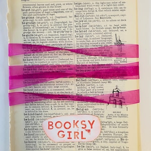 May include: A close-up of a pink and white ribbon on a page of a dictionary. The page features the words "begot", "behave", "behold", "behest", "behind", "be-hind-hand", "behold", "beholden", "behoof", "belie", "beige", "being", "Bei-rut", "be-jewel", "be-labor", "be-lated", "be-lay", "belch", "bel-dam", "be-lea-guer", "Bel-fast", "bel-fry", "Belg", "Belgian", "Belgian", "Bel-gium", "Bel-gium", "BOOKSY GIRL".
