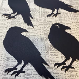 May include: Four black raven silhouettes cut from paper, arranged on a vintage book page with visible text and numbers. The ravens have detailed wing and tail shapes, with visible feet and beaks.