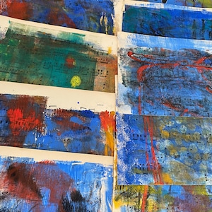 May include: A collection of abstract art pieces. Each piece features a combination of blue, red, and yellow paint strokes over vintage sheet music. The artwork has a textured appearance, with visible brushstrokes and layered colors.