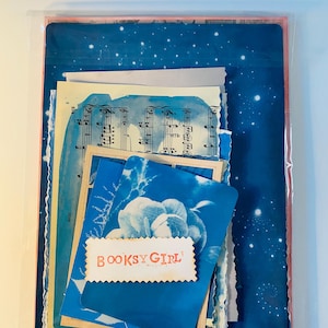 May include: A collection of ephemera, including sheet music, blue floral cards, and a card with the text "BOOKSY GIRL," packaged in a clear plastic sleeve. The items have a vintage aesthetic with a blue and white color scheme.