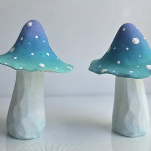 May include: Two decorative mushroom figurines. The mushroom caps are ombre, transitioning from blue to teal, with white polka dots. The stems are white with a textured, carved appearance. The mushrooms are displayed on a white surface.
