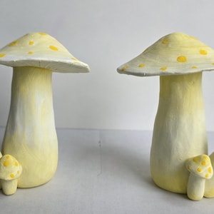 May include: Two decorative mushroom sculptures, hand-painted in pale yellow with yellow polka dots on the cap. Each mushroom has a stem and a cap. One has two smaller mushrooms attached. Suitable for home decor, these sculptures are charming.