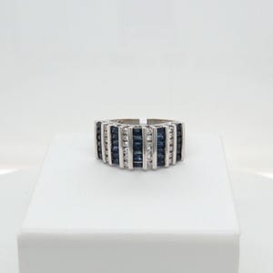 May include: A wide silver-toned band ring featuring alternating vertical channels of dark blue baguette-cut stones and clear round-cut stones. The design consists of multiple rows of these stones set between polished metal bars in a structured pattern.