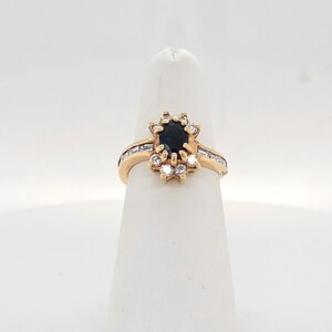 May include: A gold ring featuring a central oval-cut dark blue gemstone surrounded by a halo of small round white diamonds. The band is accented with channel-set baguette-cut diamonds along the sides, creating a detailed and structured design.