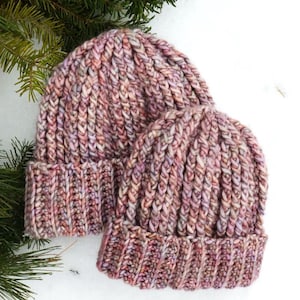 Könnte beinhalten: Zwei gestrickte Mützen in Rosa-, Lila- und Cremetönen liegen auf Schnee, mit Tannenzweigen im Hintergrund. Die Mützen haben einen gerippten Bund und ein strukturiertes, vertikales Strickmuster.