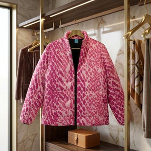 May include: A bright pink and white puffer jacket featuring a repeating snakeskin pattern, a high collar, and a black front zip closure. The jacket is displayed on a gold hanger, showcasing long sleeves and a quilted construction throughout.
