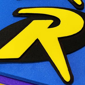May include: Two layered letter R emblems featuring a bright yellow capital letter R on top of a black oval background. The emblems are attached to a flat blue surface. The design has a bold, graphic appearance with clean edges and high contrast.