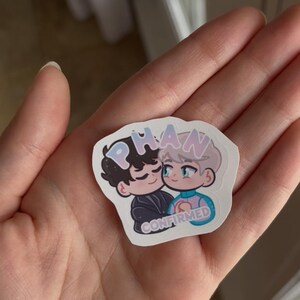 Puede incluir: Una pegatina con una ilustraci&oacute;n de dibujos animados de dos personajes abraz&aacute;ndose. La pegatina tiene la palabra "PHAN" en colores pastel en la parte superior y la palabra "CONFIRMED" en la parte inferior. Los personajes son de estilo chibi.