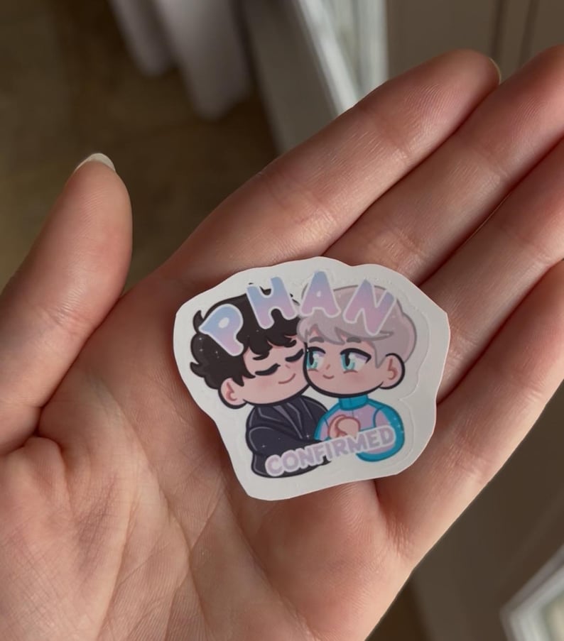 Puede incluir: Una pegatina con una ilustraci&oacute;n de dibujos animados de dos personajes abraz&aacute;ndose. La pegatina tiene la palabra "PHAN" en colores pastel en la parte superior y la palabra "CONFIRMED" en la parte inferior. Los personajes son de estilo chibi.