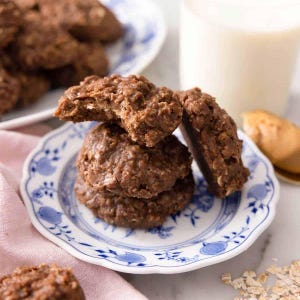 Op de afbeelding: Een stapel chocolade no-bake koekjes op een wit en blauw patroon bord. Eén koekje is gebroken en onthult de binnenkant. Een glas melk en een bord met koekjes staan op de achtergrond. De koekjes hebben een textuur.