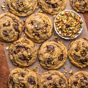 May include: Overhead shot of freshly baked chocolate chip cookies with chopped pistachios and sea salt. The cookies are golden brown with dark chocolate chunks and pistachio pieces. A small white bowl of chopped pistachios sits nearby.