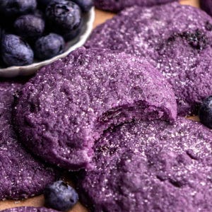 May include: Close-up of several purple blueberry cookies, dusted with sugar. One cookie has a bite taken out of it. A small white bowl filled with fresh blueberries is in the background. The cookies are on a light brown surface.