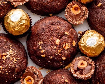 Peut inclure: Vue aérienne de desserts et bonbons au chocolat. Plusieurs pâtisseries rondes, brun foncé, sont dispersées autour de chocolats enveloppés d'or et de chocolats coupés en deux, révélant un centre de noisette. Les pâtisseries sont saupoudrées de noix hachées.