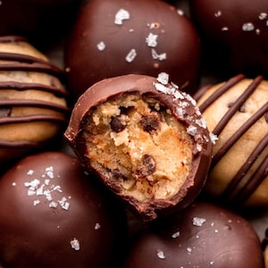 Könnte beinhalten: Nahaufnahme von schokoladenüberzogenen Keksteigkugeln. Eine Kugel ist halbiert und zeigt das Innere des Teigs mit Schokoladenstückchen. Die dunkle Schokoladenbeschichtung ist mit grobem Meersalz bestreut. Weitere Kugeln sind im Hintergrund zu sehen.