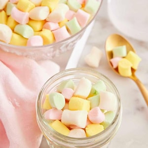 Könnte beinhalten: Nahaufnahme von bunten, pastellfarbenen Marshmallows in einem Glas und einer durchsichtigen Glasschüssel. Die Marshmallows sind gelb, rosa, grün und weiß. Ein goldener Löffel hält ein paar Marshmallows. Ein rosa Tuch ist im Hintergrund.