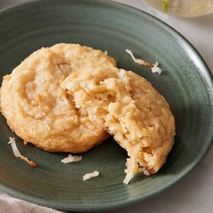 Op de afbeelding: Twee goudbruine kokoskoekjes op een donkergroen bord. Eén koekje is heel, de andere is in tweeën gebroken en toont een zacht, gestructureerd interieur. Kokosvlokken zijn verspreid.