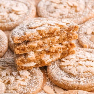 Op de afbeelding: Een stapel amandelkoekjes, bestrooid met poedersuiker, met een close-up van de textuur van de koekjes. De koekjes zijn goudbruin, met zichtbare amandelschilfers. Andere koekjes liggen verspreid rond de stapel.