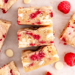 Könnte beinhalten: Draufsicht auf drei Himbeer- und weiße Schokoladen-Blondies, die übereinander gestapelt sind. Die Blondies sind hellbraun mit roten Himbeeren und weißen Schokoladenstücken. Frische Himbeeren und weiße Schokoladenchips sind verstreut.