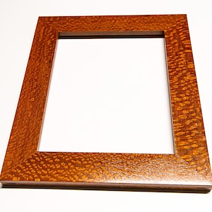 May include: A rectangular wooden picture frame with a warm, reddish-brown finish. The frame has a square opening in the center and a textured surface. The wood grain is visible, adding to the frame's natural aesthetic. The frame is angled, showcasing its depth and design.