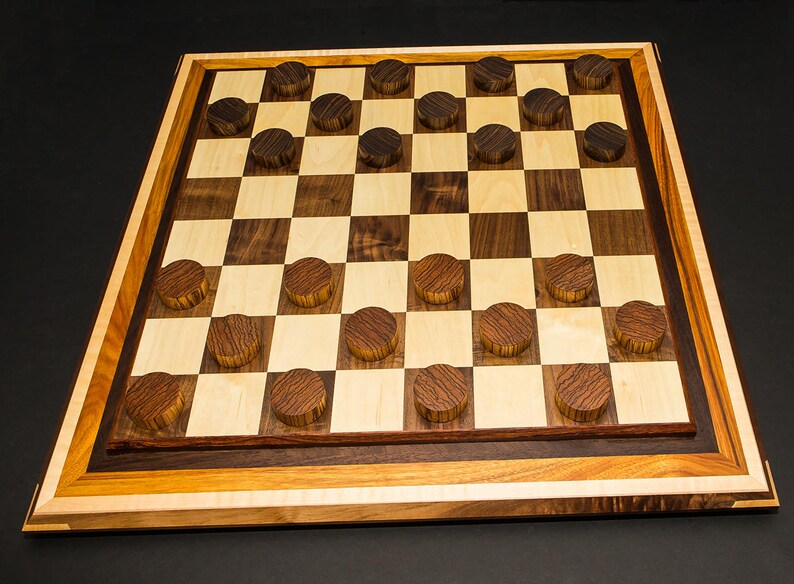 May include: A handcrafted wooden checkerboard with alternating light and dark squares. The board is framed with multiple wood tones and includes dark wooden checker pieces. The game board is set against a dark background.