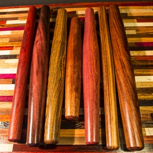 May include: Seven wooden rolling pins of various wood types and colors, arranged on a colorful wooden cutting board.