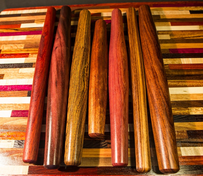 May include: Seven wooden rolling pins of various wood types and colors, arranged on a colorful wooden cutting board.