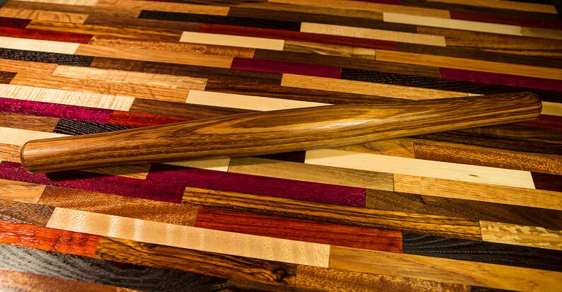 May include: A wooden rolling pin with a dark brown finish, lying on a colorful wooden cutting board made of various wood types.