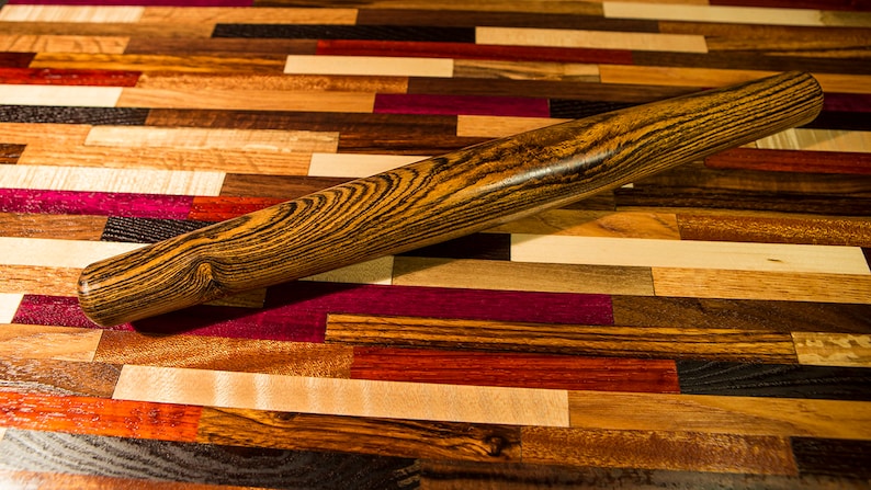 May include: A wooden rolling pin with a dark brown and black striped pattern. The rolling pin is lying on a colorful wooden surface.