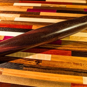 May include: A dark brown wooden rolling pin on a colorful wooden surface. The rolling pin is smooth and has a slightly rounded shape.