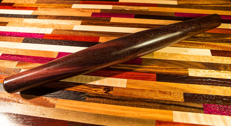 May include: A dark brown wooden rolling pin on a colorful wooden surface. The rolling pin is smooth and has a slightly rounded shape.