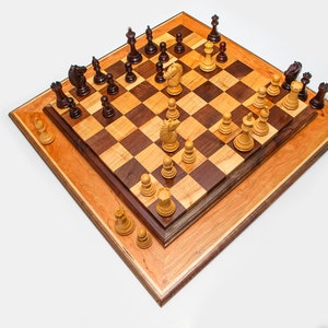 May include: A wooden chessboard with a light brown and dark brown checkered pattern. The board is set up for a game of chess with white and black pieces in place. The board is surrounded by a light brown frame.