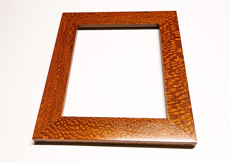May include: A rectangular wooden picture frame with a warm, reddish-brown finish. The frame has a square opening in the center and a textured surface. The wood grain is visible, adding to the frame's natural aesthetic. The frame is angled, showcasing its depth and design.