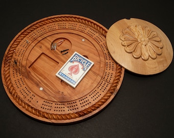 Puede incluir: Un tablero de cribbage de madera con un borde de cuerda y una tapa de madera tallada. La tapa tiene un diseño floral y el tablero tiene una carta de juego encima que dice "Bicycle Standard".