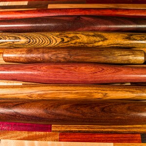 May include: A set of seven wooden rolling pins, each with a unique wood grain pattern. The rolling pins are arranged in a stack, with the darkest one at the bottom and the lightest one at the top. The rolling pins are all about the same size and shape.