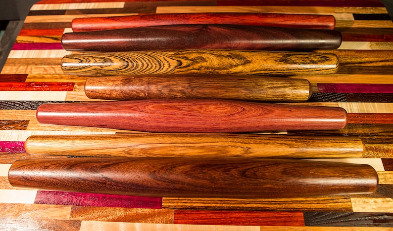 May include: A set of seven wooden rolling pins, each with a unique wood grain pattern. The rolling pins are arranged in a stack, with the darkest one at the bottom and the lightest one at the top. The rolling pins are all about the same size and shape.